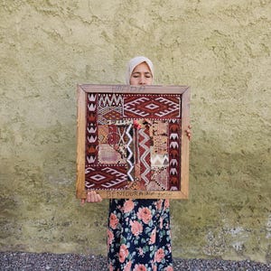 May include: A square wooden frame containing a patchwork textile with various geometric patterns in shades of red, brown, black, and cream. The top of the frame is inscribed with the word HAFIDA and the bottom with BENTOURAINE KESH.
