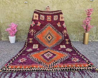 May include: A rectangular wool rug featuring a deep purple base with two large central orange diamonds outlined in black. The rug is decorated with smaller geometric patterns in green, yellow, and white, finished with a fringed edge at the bottom.