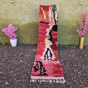 Puede incluir: Una alfombra rectangular tejida a mano con un diseño abstracto vibrante. La alfombra presenta una paleta de colores rojo, rosa, negro y crema. La alfombra tiene un borde con flecos y se exhibe al aire libre con flores rosas en el fondo.