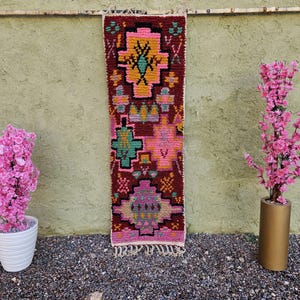 May include: A colorful, rectangular rug with a geometric pattern in shades of burgundy, pink, yellow, and teal hangs on a wall. Two pink flowering plants in white and gold pots flank the rug. The rug has a fringed edge.