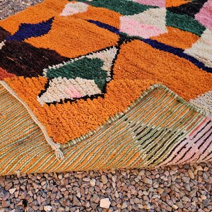 May include: A colorful, geometric patterned rug with a mix of orange, green, pink, and blue hues. The rug's design features a patchwork of shapes and textures, with a visible underside showing a striped pattern. The rug is placed on a bed of small, brown pebbles.