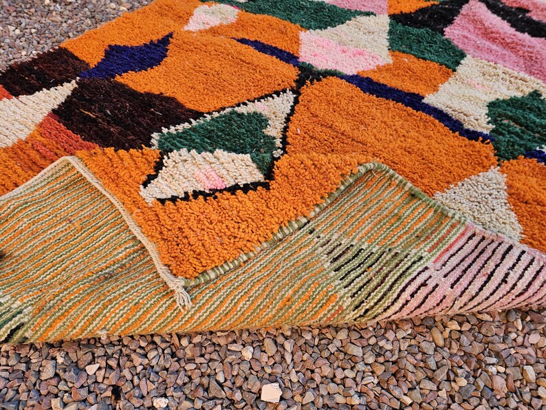 May include: A colorful, geometric patterned rug with a mix of orange, green, pink, and blue hues. The rug's design features a patchwork of shapes and textures, with a visible underside showing a striped pattern. The rug is placed on a bed of small, brown pebbles.