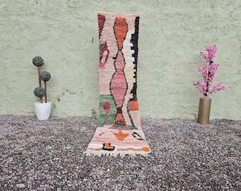 May include: A rectangular, colorful rug with a geometric design. The rug features a mix of pink, orange, green, and black shapes on a cream background. The rug is displayed vertically against a textured green wall.