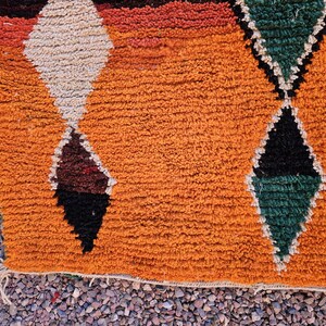 May include: Close-up of a textured orange rug with geometric diamond patterns. The diamonds are in shades of white, brown, black, and green. The rug has a fringe along the bottom edge and is likely made of wool.