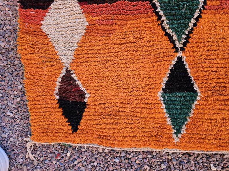 May include: Close-up of a textured orange rug with geometric diamond patterns. The diamonds are in shades of white, brown, black, and green. The rug has a fringe along the bottom edge and is likely made of wool.