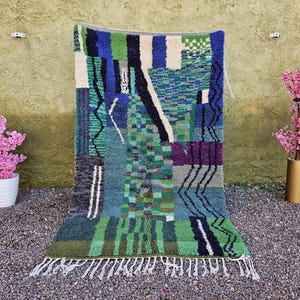 May include: A colorful, geometric rug with a patchwork design. The rug features a variety of colors, including green, blue, purple, and white. It has a fringed edge and is displayed against a textured wall.