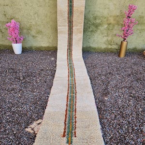 Puede incluir: Una alfombra larga y rectangular de color beige con una franja vertical central en verde, naranja y turquesa. La alfombra tiene una superficie texturizada y flecos en la parte inferior. La alfombra se exhibe al aire libre.