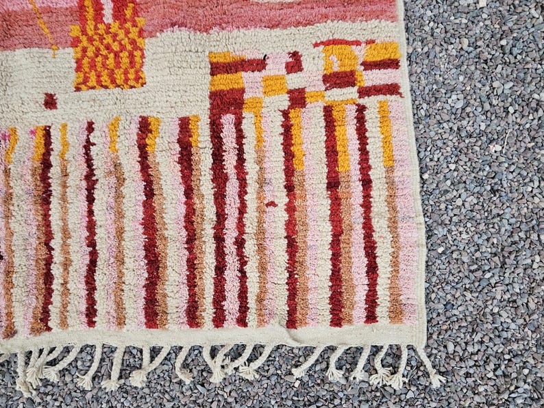 Op de afbeelding: Close-up van een handgeweven tapijt met een getextureerde, gebroken witte achtergrond. Het tapijt heeft verticale strepen in tinten rood, roze en geel, samen met geometrische vormen. De onderkant heeft een franje.