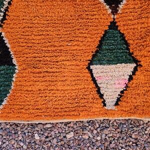 May include: Close-up of a textured orange rug with diamond patterns. The diamonds are outlined in black and filled with green, beige, and pink. The rug has a fringe along the bottom edge and is set against a background of small pebbles.