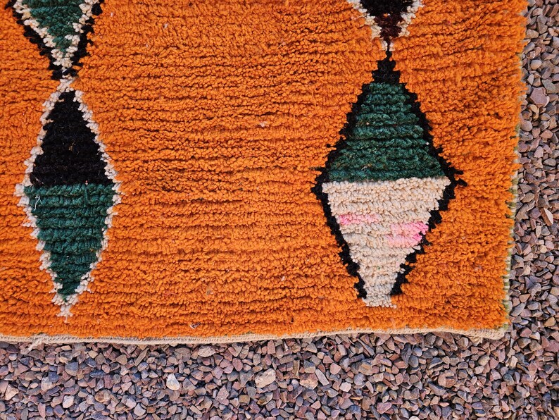 May include: Close-up of a textured orange rug with diamond patterns. The diamonds are outlined in black and filled with green, beige, and pink. The rug has a fringe along the bottom edge and is set against a background of small pebbles.