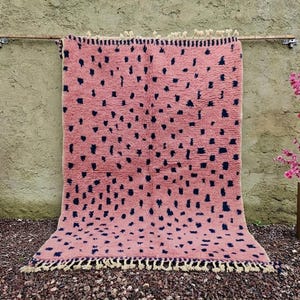 May include: A pink rug with a dark blue spot pattern hangs against a textured wall. The rug has a fringe along the bottom edge. Two potted plants with pink flowers flank the rug, adding a pop of color to the scene.