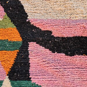 May include: Close-up of a colorful, textured rug with geometric shapes. The rug features a combination of pink, black, orange, green, and beige hues. The design includes a large black shape and several geometric patterns.
