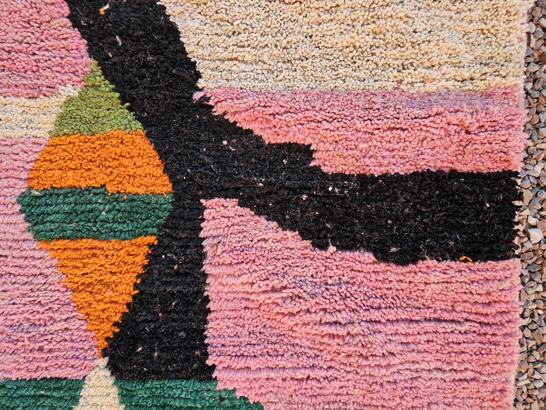 May include: Close-up of a colorful, textured rug with geometric shapes. The rug features a combination of pink, black, orange, green, and beige hues. The design includes a large black shape and several geometric patterns.