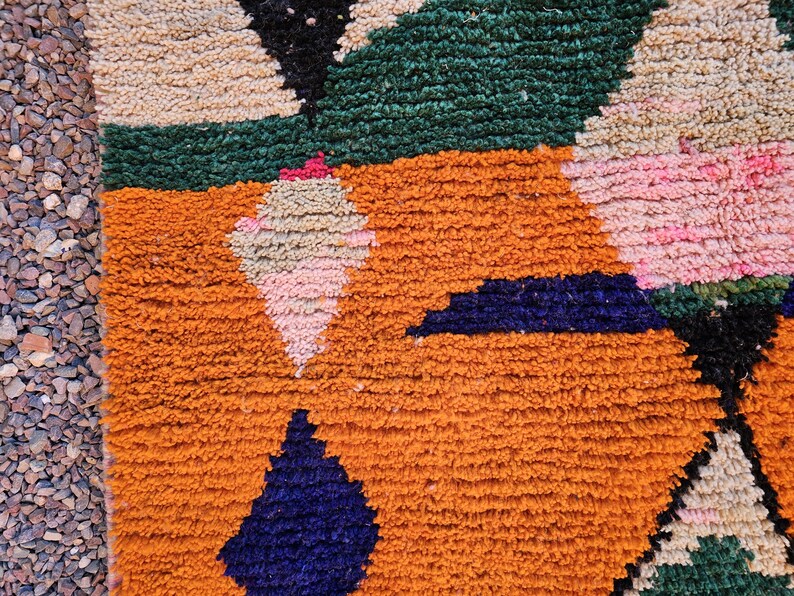 May include: Close-up of a colorful, textured rug with geometric patterns. The rug features orange, green, black, pink, and blue shapes. The rug's design is a mix of triangles and diamond shapes, creating a vibrant and eye-catching textile.