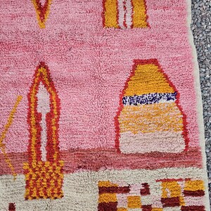 Op de afbeelding: Roze en beige tapijt met geometrische ontwerpen in geel, oranje en rood. Het tapijt heeft een getextureerd, geweven uiterlijk. Het ontwerp bevat vormen die lijken op gebouwen en abstracte vormen.