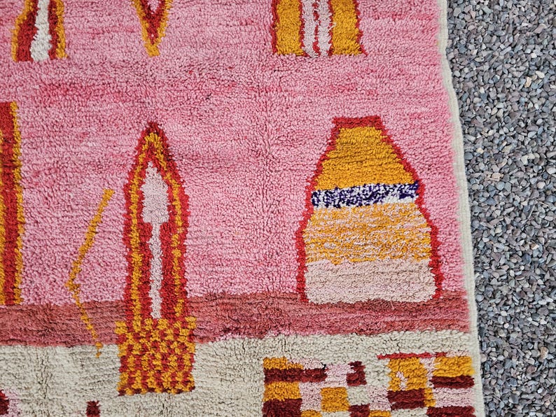 Op de afbeelding: Roze en beige tapijt met geometrische ontwerpen in geel, oranje en rood. Het tapijt heeft een getextureerd, geweven uiterlijk. Het ontwerp bevat vormen die lijken op gebouwen en abstracte vormen.