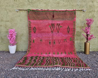 May include: A vibrant pink Moroccan rug with green and brown geometric patterns. The rug features a central design of diamonds and stylized shapes, with a fringed edge. The rug is displayed against a textured wall, with pink flowers in pots on either side.