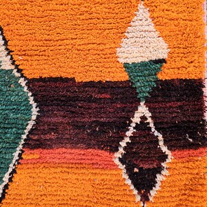 May include: Close-up of a vibrant, textured rug with a geometric diamond pattern. The rug features a warm orange background with accents of green, white, and dark red. The design is bold and eye-catching.