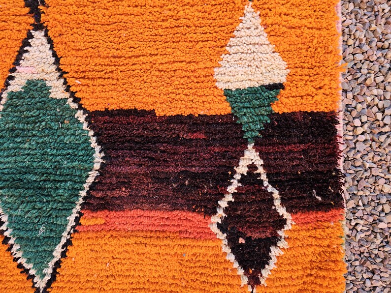 May include: Close-up of a vibrant, textured rug with a geometric diamond pattern. The rug features a warm orange background with accents of green, white, and dark red. The design is bold and eye-catching.