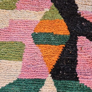 May include: Close-up of a colorful, textured rug with a geometric design. The rug features a combination of pink, green, orange, black, and beige rectangular and triangular shapes. The rug has a plush, woven texture.