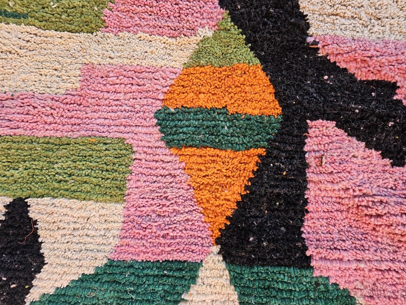 May include: Close-up of a colorful, textured rug with a geometric design. The rug features a combination of pink, green, orange, black, and beige rectangular and triangular shapes. The rug has a plush, woven texture.