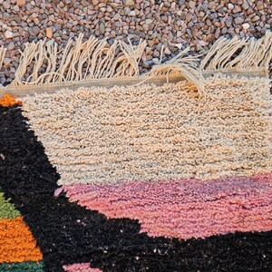 May include: Close-up of a colorful, textured rug with a geometric design. The rug features a mix of colors including black, pink, orange, green, and beige. The edge has a fringe detail.