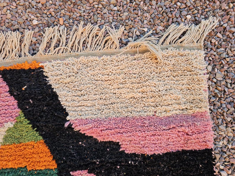 May include: Close-up of a colorful, textured rug with a geometric design. The rug features a mix of colors including black, pink, orange, green, and beige. The edge has a fringe detail.