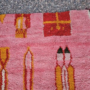Op de afbeelding: Roze tapijt met geometrische patronen in rood, geel en wit. Het tapijt heeft herhalende vormen, waaronder rechthoeken en gestileerde figuren. De textuur lijkt zacht en pluche, met een close-up die de geweven details benadrukt.
