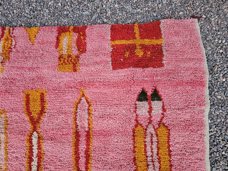 Op de afbeelding: Roze tapijt met geometrische patronen in rood, geel en wit. Het tapijt heeft herhalende vormen, waaronder rechthoeken en gestileerde figuren. De textuur lijkt zacht en pluche, met een close-up die de geweven details benadrukt.