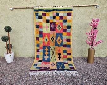 Può includere: Un tappeto rettangolare colorato con un motivo geometrico. Il tappeto presenta quadrati in vari colori, tra cui giallo, blu, viola e rosa. Ha una frangia bianca in basso ed è esposto contro un muro neutro.