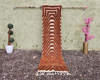 May include: A brown and white geometric patterned rug with a fringed edge hangs against a textured green wall. A potted topiary and a gold vase with pink flowers flank the rug. The rug's design features repeating chevrons.
