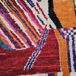 May include: Close-up of a colorful, geometric rug with a textured surface. The rug features a mix of vibrant colors, including red, orange, pink, purple, and white. The design includes diagonal stripes and rectangular shapes.
