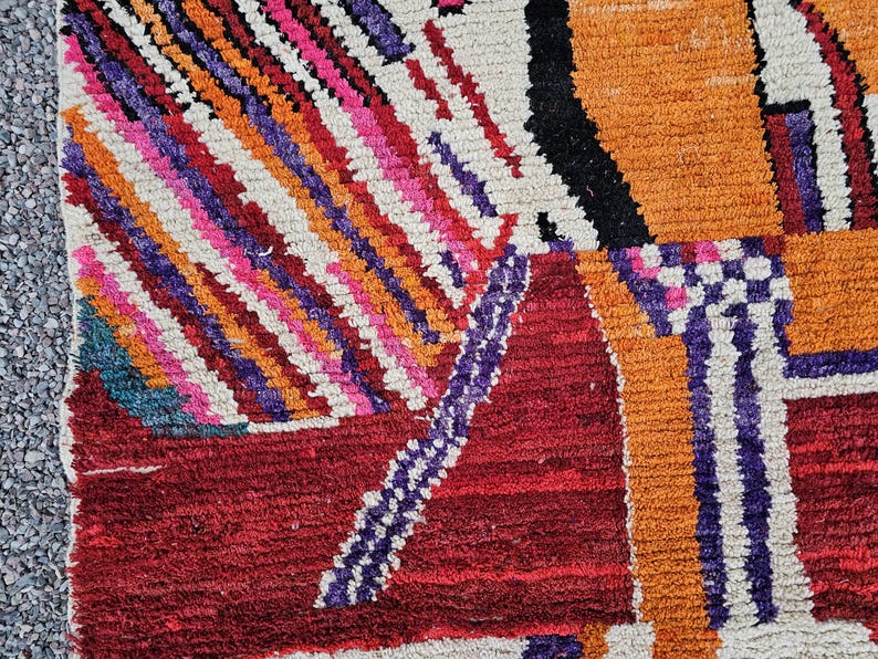 May include: Close-up of a colorful, geometric rug with a textured surface. The rug features a mix of vibrant colors, including red, orange, pink, purple, and white. The design includes diagonal stripes and rectangular shapes.