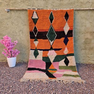 May include: A vibrant, hand-woven rug with an orange background and geometric diamond patterns in green, white, and black. The rug is displayed against a textured wall, with pink flowers in a white pot and a gold vase on either side.