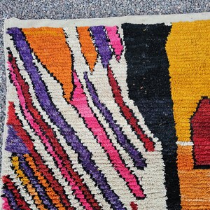 May include: Close-up of a colorful rug with a geometric pattern. The rug features a mix of vibrant colors, including orange, purple, pink, red, black, and yellow. The design includes diagonal stripes and a central shape.