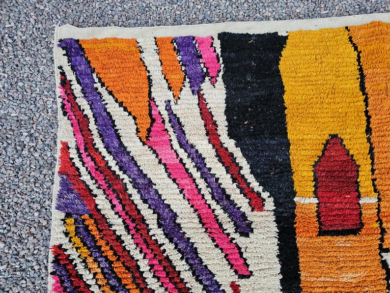 May include: Close-up of a colorful rug with a geometric pattern. The rug features a mix of vibrant colors, including orange, purple, pink, red, black, and yellow. The design includes diagonal stripes and a central shape.