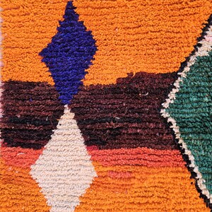 May include: Close-up of a vibrant, textured rug with a geometric design. The rug features a warm orange background with a large blue diamond shape, a white diamond, and a green diamond. The rug has a shaggy texture.