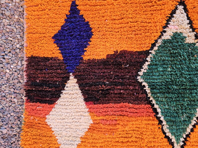 May include: Close-up of a vibrant, textured rug with a geometric design. The rug features a warm orange background with a large blue diamond shape, a white diamond, and a green diamond. The rug has a shaggy texture.