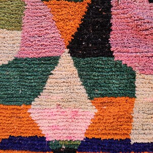 May include: Close-up of a colorful, textured rug with a geometric pattern. The rug features a mix of colors including pink, orange, green, black, and cream. The design includes diamond and rectangular shapes.