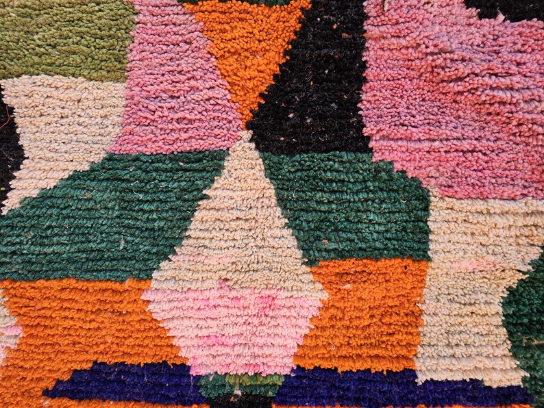 May include: Close-up of a colorful, textured rug with a geometric pattern. The rug features a mix of colors including pink, orange, green, black, and cream. The design includes diamond and rectangular shapes.