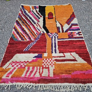 May include: A rectangular, hand-woven rug with a vibrant, abstract design. The rug features a mix of colors including red, orange, yellow, white, and purple. The design includes geometric shapes and patterns, with a fringed edge.