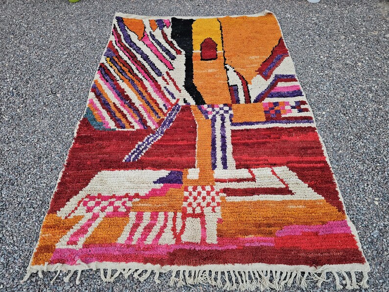 May include: A rectangular, hand-woven rug with a vibrant, abstract design. The rug features a mix of colors including red, orange, yellow, white, and purple. The design includes geometric shapes and patterns, with a fringed edge.