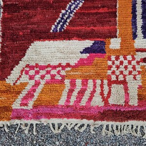 May include: A close-up of a colorful, textured rug with a geometric design. The rug features a mix of colors, including red, orange, pink, and white. The design includes stripes, squares, and other shapes. The rug has a fringe along the bottom edge.