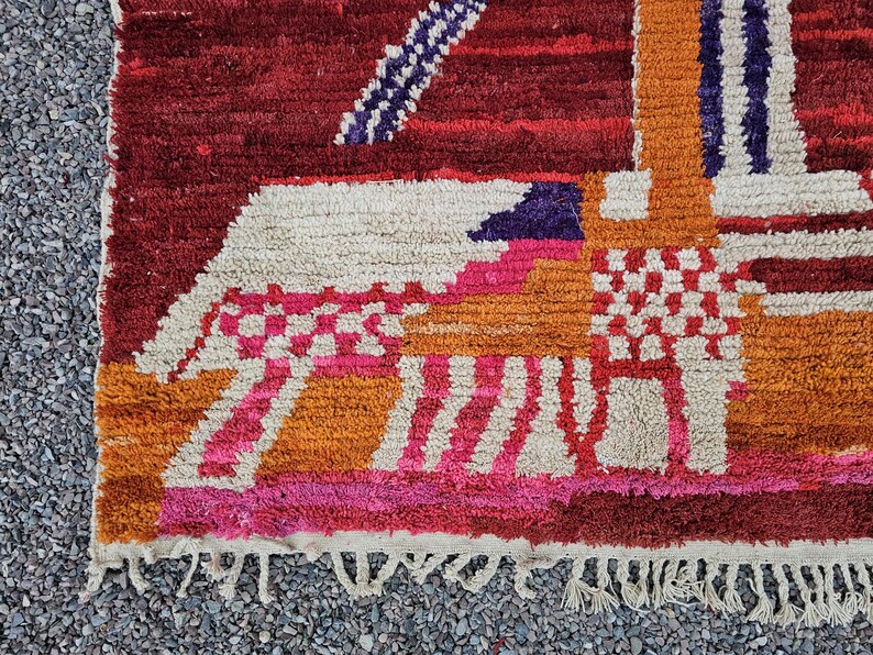 May include: A close-up of a colorful, textured rug with a geometric design. The rug features a mix of colors, including red, orange, pink, and white. The design includes stripes, squares, and other shapes. The rug has a fringe along the bottom edge.