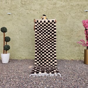 May include: A black and white checkered rug with a fringed edge is held up against a textured green wall. A potted topiary and a vase with pink flowers flank the rug. The rug's pattern is a repeating grid of black squares on a white background.