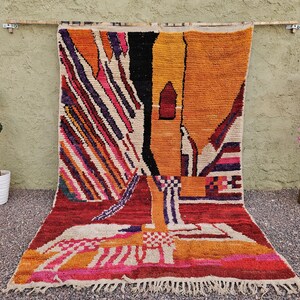 May include: A colorful, abstract Moroccan rug with a geometric design. The rug features a vibrant palette of red, orange, yellow, black, and white, with fringe along the bottom edge. The rug is displayed against a neutral background.