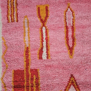 Op de afbeelding: Roze, getextureerd tapijt met geometrische ontwerpen in rood, geel en wit. Het tapijt heeft abstracte vormen, waaronder rechthoeken, driehoeken en langwerpige vormen, waardoor een levendig en artistiek patroon ontstaat. Het ontwerp van het tapijt voegt kleur en textuur toe.