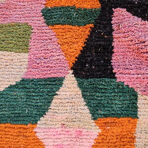 May include: Close-up of a colorful, textured rug with geometric shapes. The rug features a mix of pink, orange, green, black, and cream colors. The design includes triangles and rectangular shapes, creating a vibrant and eye-catching pattern.