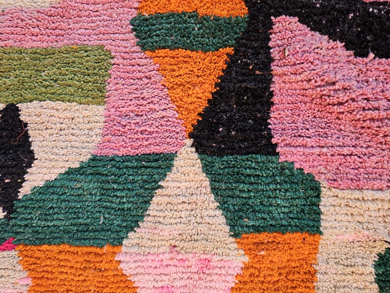 May include: Close-up of a colorful, textured rug with geometric shapes. The rug features a mix of pink, orange, green, black, and cream colors. The design includes triangles and rectangular shapes, creating a vibrant and eye-catching pattern.