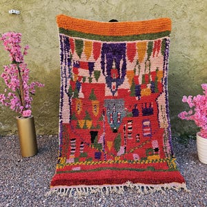 Puede incluir: Una alfombra tejida a mano de colores vibrantes con una base roja y intrincados patrones geométricos en tonos naranja, morado, verde y rosa. La alfombra tiene un borde con flecos y se exhibe contra una pared neutra, con plantas de flores rosas a ambos lados.